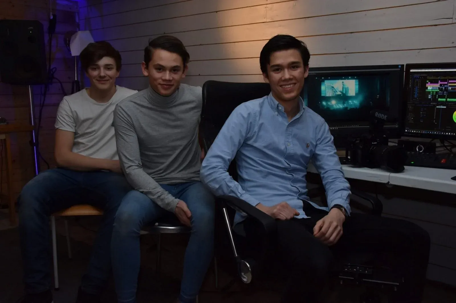 Three young men in a small video studio. One sits in an office chair in a light blue shirt, the other two on chairs beside him, in a grey turtleneck and a white T-shirt. Behind them are two screens with video editing and a camera with a microphone.