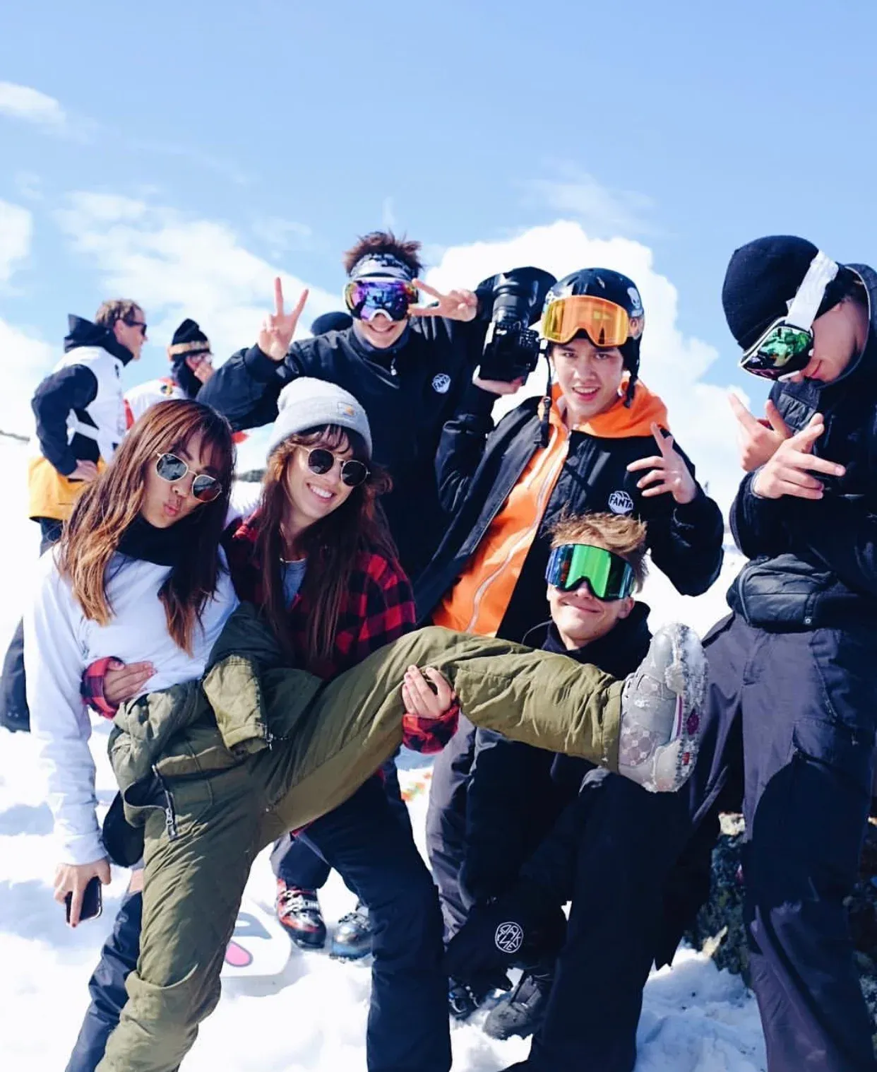 A group of young people in ski clothes in the snow in Hemsedal under a blue sky. Several pose in sunglasses and goggles, one holds a video camera and wears a Fanta jacket, and two of them carry a third between them.