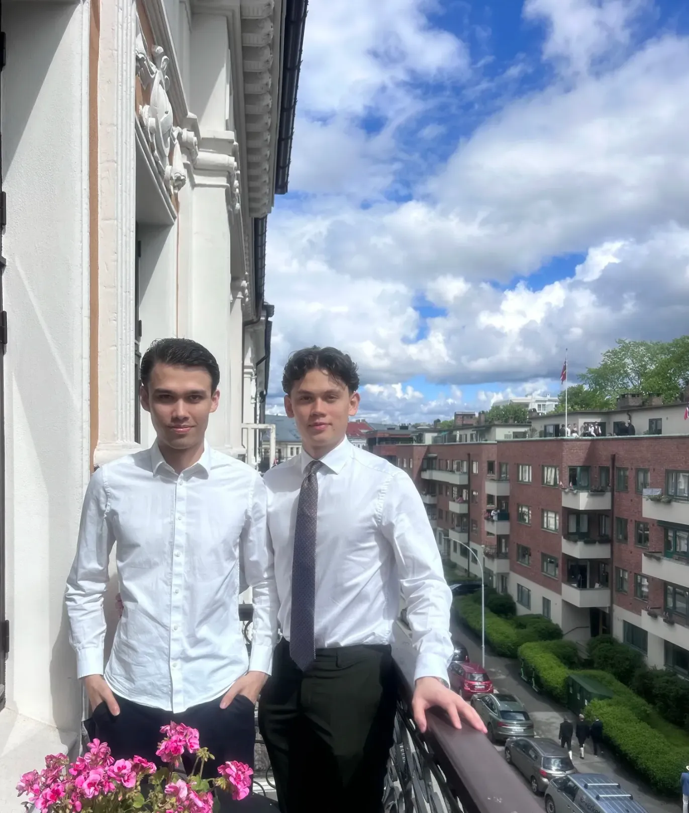 Two brothers in white shirts on a balcony in Oslo, one in a tie, with pink geraniums in front and blue sky and a Norwegian flag on the rooftops behind them